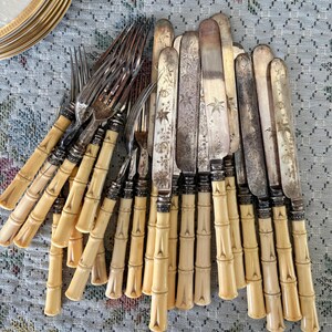 Pode incluir: Uma cole&ccedil;&atilde;o de talheres antigos, incluindo garfos e facas. Os garfos t&ecirc;m cabos em estilo bambu com detalhes prateados. As facas apresentam gravuras florais nas l&acirc;minas. Os talheres est&atilde;o dispostos sobre uma toalha de mesa estampada.
