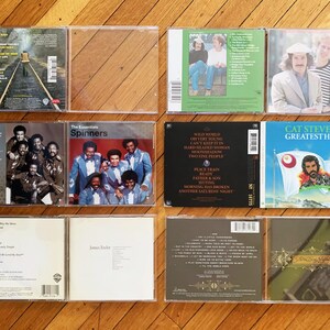May include: A collection of ten compact discs in clear plastic cases. The cases are arranged in two rows of five. The discs feature a variety of artists and genres, including The Spinners, Cat Stevens, and James Taylor. The titles of the albums are visible on the cases.