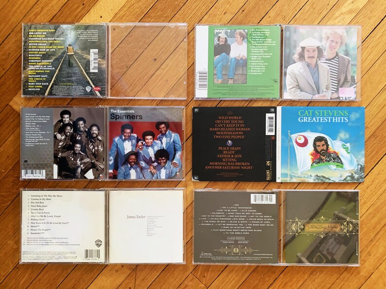 May include: A collection of ten compact discs in clear plastic cases. The cases are arranged in two rows of five. The discs feature a variety of artists and genres, including The Spinners, Cat Stevens, and James Taylor. The titles of the albums are visible on the cases.