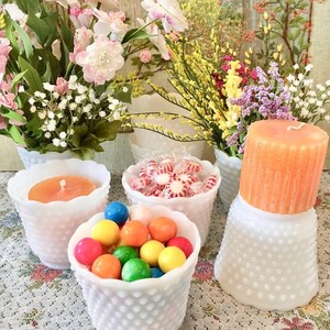 May include: A collection of white hobnail glass containers filled with colorful candies and a candle. The containers hold gumballs, peppermints, and a large orange candle. Floral arrangements in white vases are in the background.