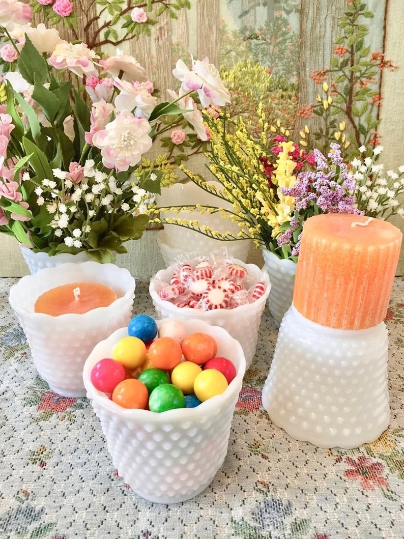 May include: A collection of white hobnail glass containers filled with colorful candies and a candle. The containers hold gumballs, peppermints, and a large orange candle. Floral arrangements in white vases are in the background.