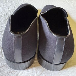 Puede incluir: Un par de zapatos negros sin cordones con punta redonda y tac&oacute;n bajo. Los zapatos tienen un acabado liso y mate con un ribete plateado contrastante alrededor de la abertura y el tal&oacute;n. Los zapatos son de tela.