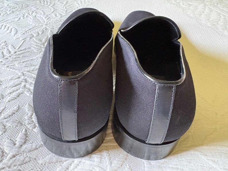 Puede incluir: Un par de zapatos negros sin cordones con punta redonda y tac&oacute;n bajo. Los zapatos tienen un acabado liso y mate con un ribete plateado contrastante alrededor de la abertura y el tal&oacute;n. Los zapatos son de tela.