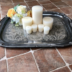 May include: A silver tray with ornate detailing holds a collection of white candles. The candles are arranged in a pyramid shape, with a larger candle at the top and smaller candles surrounding it. A bouquet of yellow and white flowers is placed on the tray.