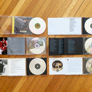 May include: A collection of eight CDs in clear plastic cases, with black and white album covers. The CDs are arranged in two rows of four. The album covers feature a group of people in red and white clothing, a man in a black leather jacket, a group of people in white clothing, and a group of people in black clothing. The CDs are labeled with the titles "The Jackson 5", "Going Back to My Roots", "The Best of The Jackson 5", "The Jackson 5: The Ultimate Collection", "The Jackson 5: The Essential Collection", "The Jackson 5: The Very Best of", "The Jackson 5: The Complete Motown Singles", and "The Jackson 5: The Complete Albums".