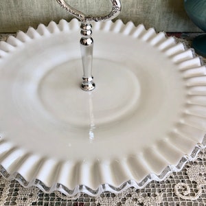 Puede incluir: Un soporte para pastel de vidrio blanco lechoso con un asa plateada. El soporte tiene un borde festoneado y un centro elevado.