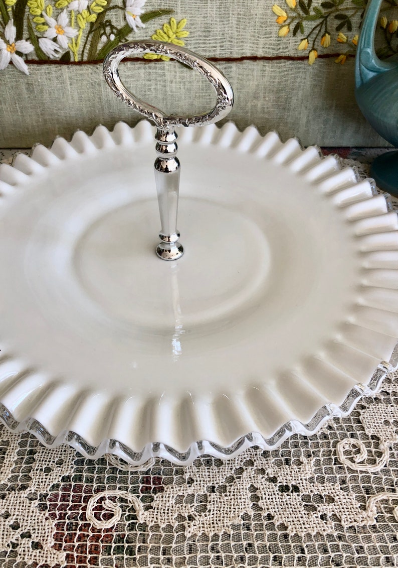 Puede incluir: Un soporte para pastel de vidrio blanco lechoso con un asa plateada. El soporte tiene un borde festoneado y un centro elevado.
