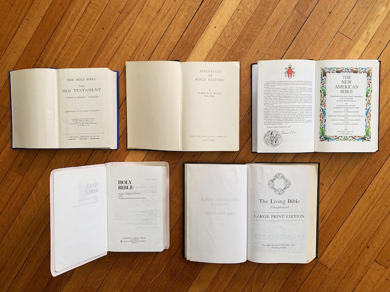Op de afbeelding: Een bovenaanzicht van zes open boeken op een houten oppervlak. De boeken tonen titels als "Oude Testament", "Essentials of Bible History" en "The New American Bible". De pagina's zijn wit met zwarte tekst.