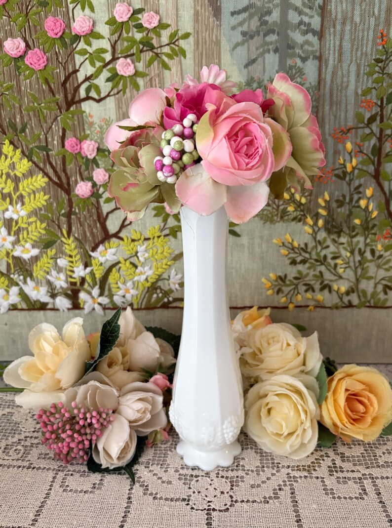 Peut inclure: Un vase blanc de style vintage rempli de fleurs artificielles roses et vertes. D'autres fleurs dans les tons de rose, cr&egrave;me et jaune sont dispos&eacute;es autour du vase. L'arri&egrave;re-plan pr&eacute;sente une tapisserie avec des motifs floraux et d'arbres.