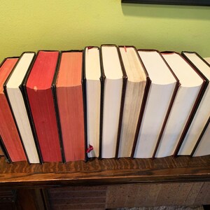 May include: A row of ten hardback books with red and white spines. The books are arranged in a line on a wooden surface.