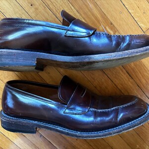 May include: A pair of dark brown leather penny loafers featuring a classic slip-on design with a rounded toe, a stitched moccasin-style seam across the vamp, a leather strap with a horizontal cutout, and a stacked wooden heel with a black sole.