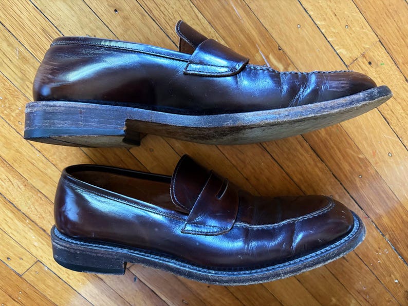 May include: A pair of dark brown leather penny loafers featuring a classic slip-on design with a rounded toe, a stitched moccasin-style seam across the vamp, a leather strap with a horizontal cutout, and a stacked wooden heel with a black sole.