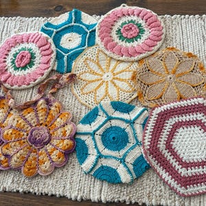 May include: A collection of handmade crochet items in various shapes and colors. Includes round, flower-shaped, and geometric designs in shades of pink, blue, yellow, and orange. The items are displayed on a textured, neutral-colored surface.