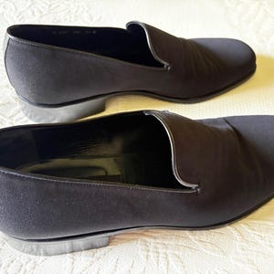 Puede incluir: Un par de zapatos negros sin cordones. Los zapatos tienen una punta redondeada y un tac&oacute;n bajo. La parte superior es de tela lisa y oscura. Los zapatos se exhiben sobre una superficie blanca.