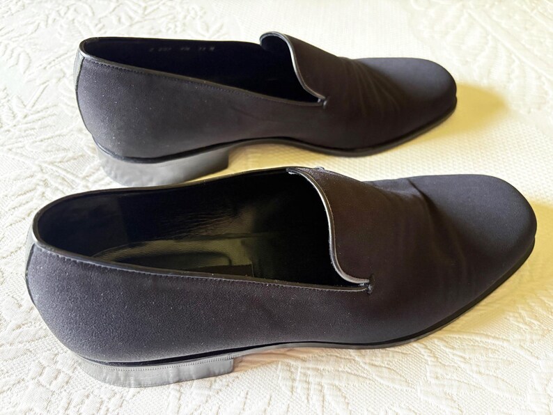 Puede incluir: Un par de zapatos negros sin cordones. Los zapatos tienen una punta redondeada y un tac&oacute;n bajo. La parte superior es de tela lisa y oscura. Los zapatos se exhiben sobre una superficie blanca.