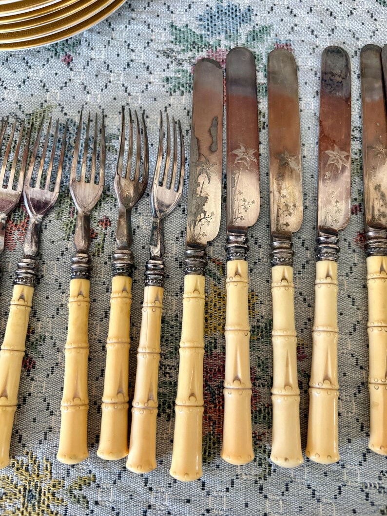 Pode incluir: Uma cole&ccedil;&atilde;o de talheres antigos, incluindo garfos e facas. Os garfos t&ecirc;m dentes prateados e cabos em estilo bambu. As facas t&ecirc;m l&acirc;minas gravadas e cabos a condizer em estilo bambu. Os talheres est&atilde;o dispostos sobre uma toalha de mesa com padr&atilde;o floral.