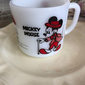 May include: White ceramic mug with a red and black illustration of Mickey Mouse wearing a red hat and a red and white striped jacket. The mug says "Mickey Mouse" in black lettering.