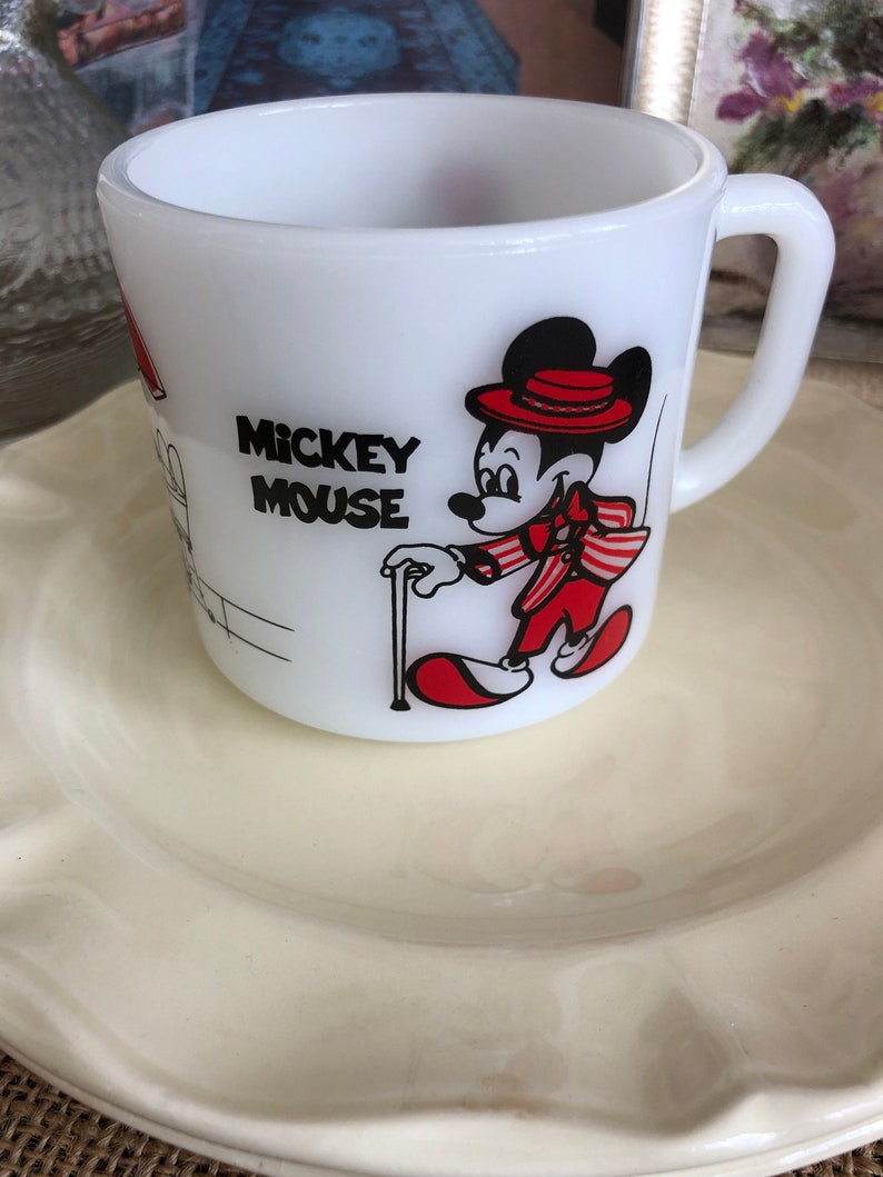 May include: White ceramic mug with a red and black illustration of Mickey Mouse wearing a red hat and a red and white striped jacket. The mug says "Mickey Mouse" in black lettering.