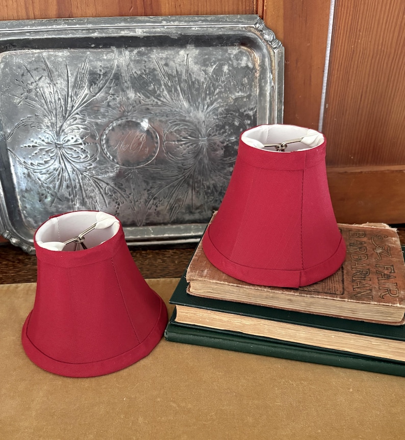 Op de afbeelding: Twee rode lampenkappen met witte binnenkant staan op een stapel oude boeken. De lampenkappen zijn dieprood van kleur en hebben een klassieke belvorm. Op de achtergrond staat een decoratieve zilveren dienblad.
