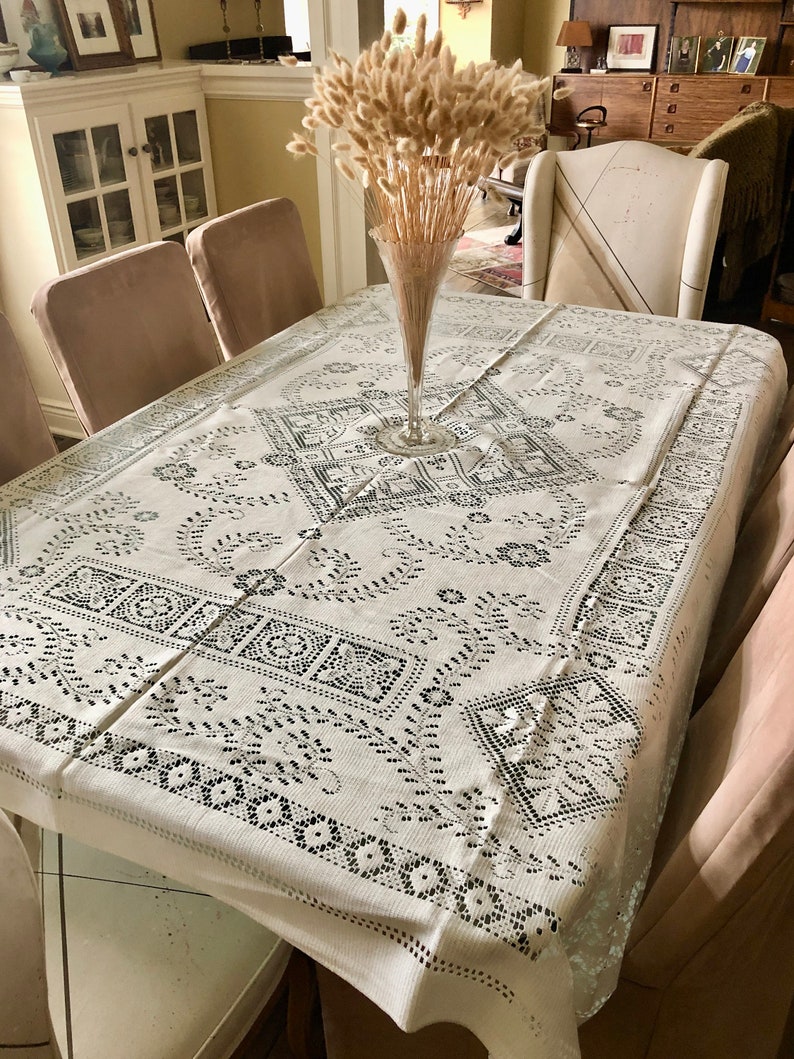 Peut inclure: Une nappe blanche avec un motif de dentelle noire, dress&eacute;e pour un d&icirc;ner. Un vase avec des fleurs s&eacute;ch&eacute;es beiges se trouve au centre de la table.