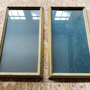 May include: Two gold-colored picture frames with ornate detailing. The frames are empty and have a glass front.