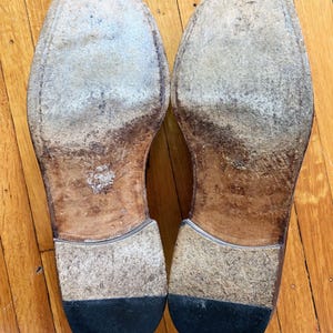 May include: A pair of worn leather shoe soles with visible scuffing and signs of use. The soles feature a tan leather base, a stacked heel construction, and a black rubber heel cap. The size 8.5 is stamped into the leather on the right sole.