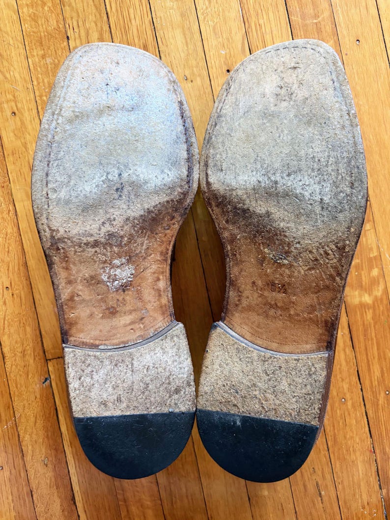 May include: A pair of worn leather shoe soles with visible scuffing and signs of use. The soles feature a tan leather base, a stacked heel construction, and a black rubber heel cap. The size 8.5 is stamped into the leather on the right sole.