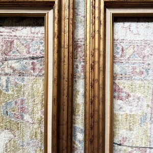May include: Two rectangular picture frames featuring a metallic gold finish with dark brown distressed detailing. Each frame has a multi-tiered moulding profile and a light beige fabric-textured inner border lining the interior edge of the frame.