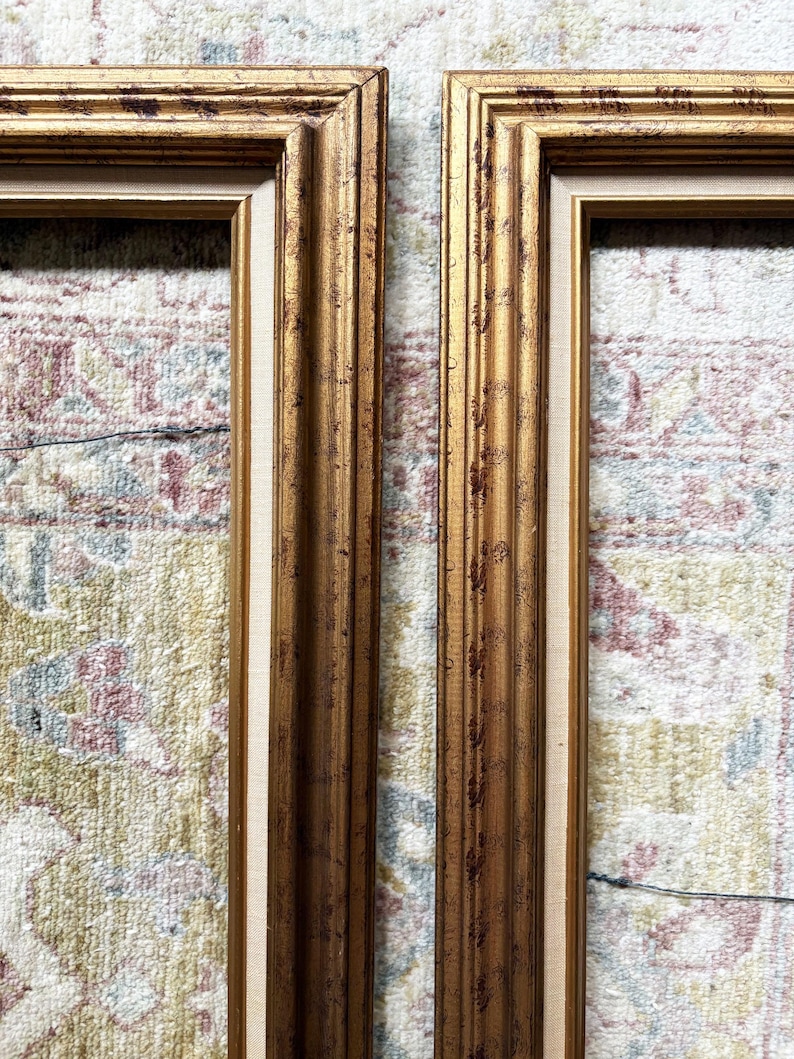May include: Two rectangular picture frames featuring a metallic gold finish with dark brown distressed detailing. Each frame has a multi-tiered moulding profile and a light beige fabric-textured inner border lining the interior edge of the frame.