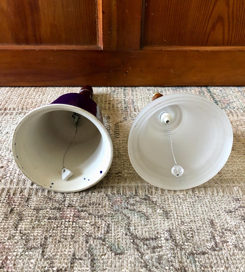 May include: Two ceramic bells, one with a blue and white glaze and the other with a frosted white glaze. Both bells have a wooden handle and a metal clapper.