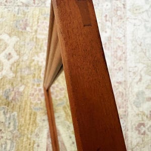 May include: Close-up of a wooden frame, likely for a mirror or artwork. The frame is a warm, reddish-brown color, with visible wood grain and joinery details. The frame is angled, showing its depth and construction.