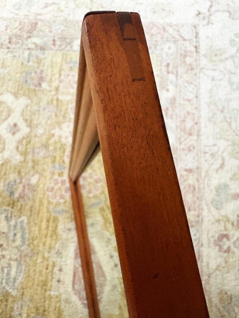 May include: Close-up of a wooden frame, likely for a mirror or artwork. The frame is a warm, reddish-brown color, with visible wood grain and joinery details. The frame is angled, showing its depth and construction.