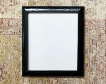May include: A black wood picture frame with a white mat and a white background. The frame is on a patterned rug.
