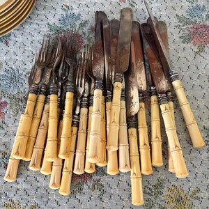 Pode incluir: Uma cole&ccedil;&atilde;o de talheres antigos, incluindo garfos e facas. As pegas t&ecirc;m um design de bambu na cor creme, e as l&acirc;minas e dentes s&atilde;o prateados. Os talheres est&atilde;o dispostos sobre uma toalha de mesa estampada.