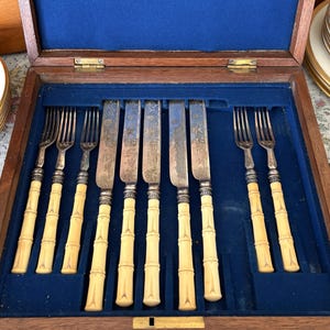 Pode incluir: Um conjunto de talheres antigos numa caixa de madeira com forro de veludo azul. O conjunto inclui garfos e facas com cabos creme em estilo bambu e l&acirc;minas prateadas. A caixa est&aacute; aberta, revelando a disposi&ccedil;&atilde;o dos utens&iacute;lios.