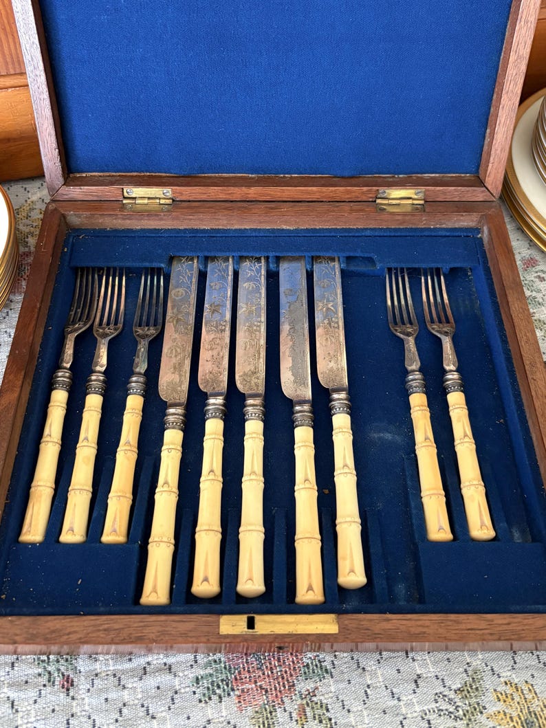 Pode incluir: Um conjunto de talheres antigos numa caixa de madeira com forro de veludo azul. O conjunto inclui garfos e facas com cabos creme em estilo bambu e l&acirc;minas prateadas. A caixa est&aacute; aberta, revelando a disposi&ccedil;&atilde;o dos utens&iacute;lios.