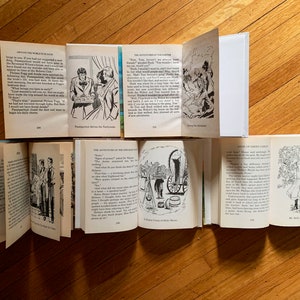 May include: A collection of vintage children's books with black and white illustrations. The books are open to different pages, showing various scenes and text. The books are on a wooden surface.