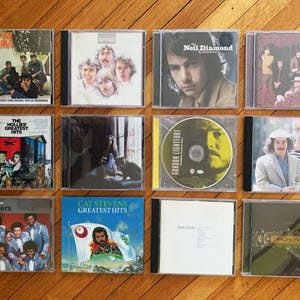 May include: A collection of 10 compact discs (CDs) featuring various artists, including The Animals, The Hollies, Neil Diamond, Gordon Lightfoot, Cat Stevens, and The Spinners. The CDs are displayed in a grid pattern on a wooden surface.