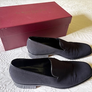 Puede incluir: Un par de zapatos negros sin cordones con tac&oacute;n bajo, junto a una caja burdeos. Los zapatos tienen un acabado liso y mate y un dise&ntilde;o cl&aacute;sico. La caja tiene una forma rectangular simple.