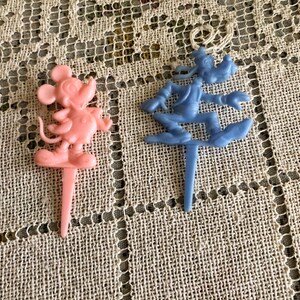May include: Two plastic cake toppers, one pink and one blue. The pink topper features Mickey Mouse, while the blue topper features Goofy.