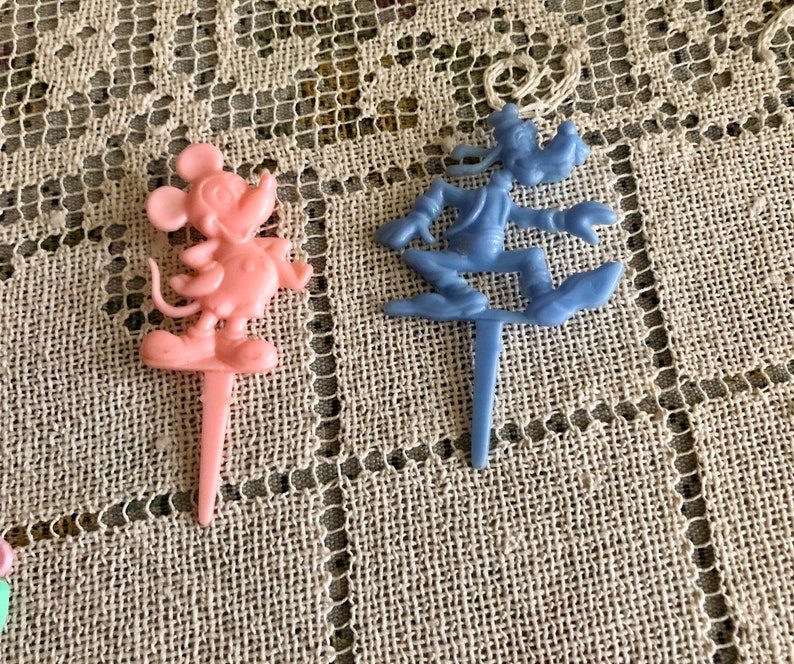 May include: Two plastic cake toppers, one pink and one blue. The pink topper features Mickey Mouse, while the blue topper features Goofy.