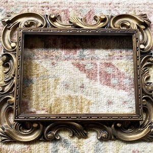 May include: A gold-colored ornate picture frame with a detailed, intricate design. The frame is made of wood and has a rectangular shape.