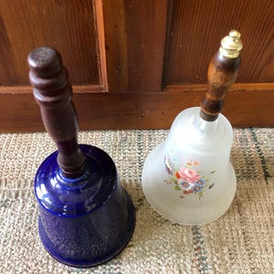 May include: Two decorative bells with wooden handles. One bell is blue with a gold trim and the other is white with a floral design.