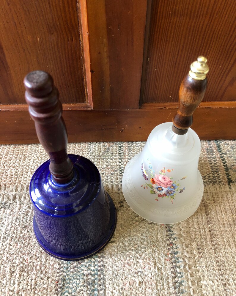 May include: Two decorative bells with wooden handles. One bell is blue with a gold trim and the other is white with a floral design.