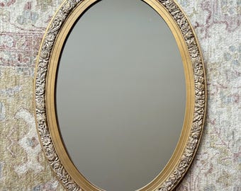 May include: An oval wall mirror featuring a gold-toned frame with an intricate, raised floral and leaf relief pattern. The mirror has a smooth, reflective glass surface surrounded by a plain inner border before the decorative outer edge.