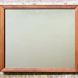May include: Rectangular mirror with a wooden frame. The frame is a warm brown color, and the mirror's surface reflects a soft, neutral hue. The mirror is a decorative item, suitable for home decor.