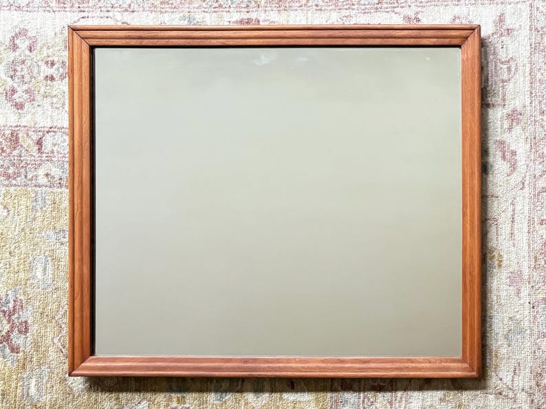 May include: Rectangular mirror with a wooden frame. The frame is a warm brown color, and the mirror's surface reflects a soft, neutral hue. The mirror is a decorative item, suitable for home decor.