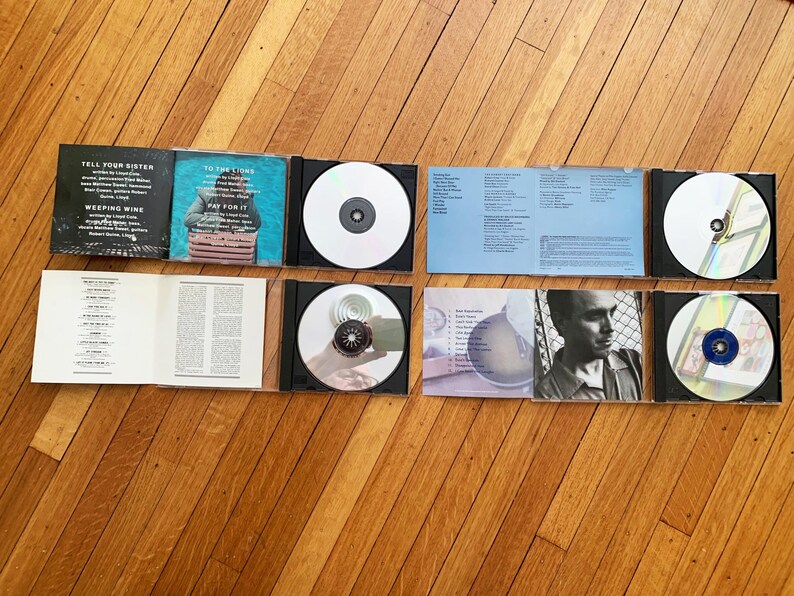 May include: A set of six CDs with accompanying booklets. The CDs are silver with various designs. The booklets are blue, white, and black with text. The text on the booklets includes the titles "Tell Your Sister", "Weeping Wine", "To The Lions", "Pay For It", and "The New Recordings".