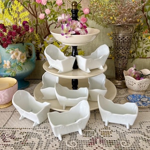 May include: A tiered serving tray with a black and gold handle, holding several small, white porcelain bathtubs. The bathtubs are arranged on cream-colored plates. The background includes floral decorations and a lace tablecloth.