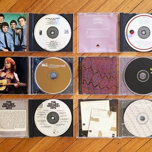 May include: A collection of nine compact discs in jewel cases. The discs feature various artists, including The Hollies, Neil Diamond, and The Guess Who. The cases are arranged in three rows of three, with the discs facing the viewer. The cases are all black with clear plastic inserts.